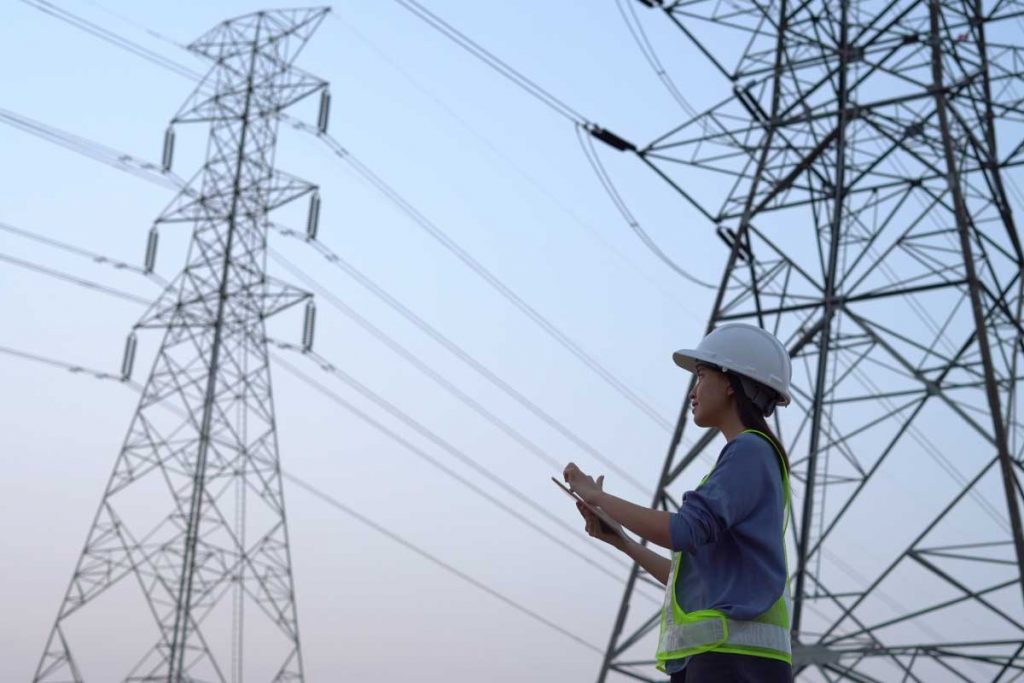 Field worker wearing a safety helmet and vest, inspecting power transmission lines at dusk with a digital tablet in hand, against a backdrop of towering pylons and a clear sky.