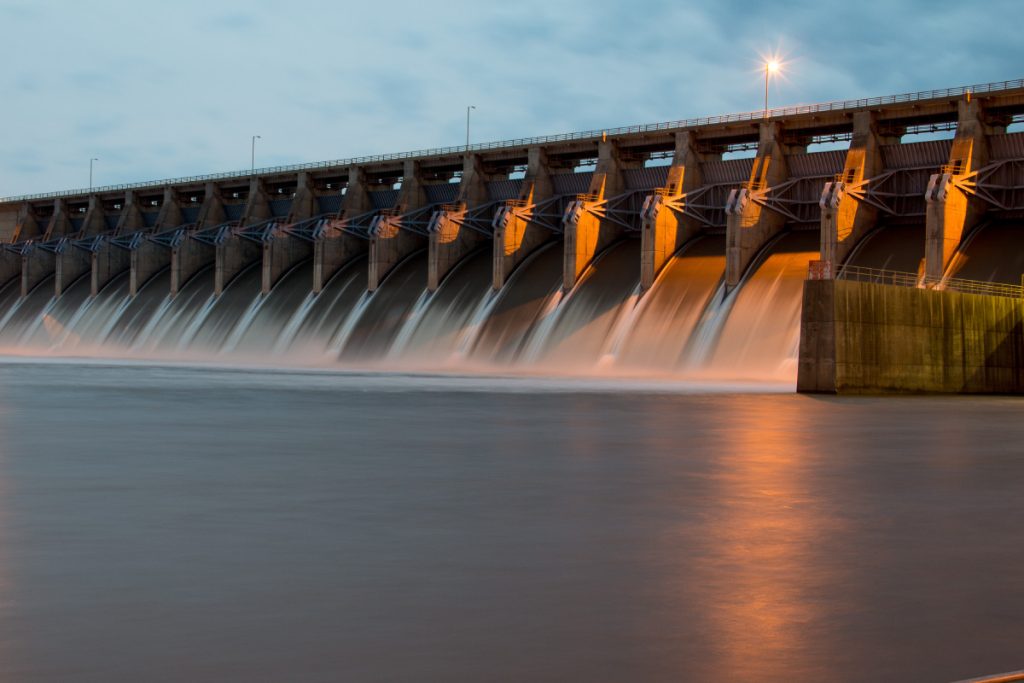 A large dam with multiple spillways releasing water, illuminated by warm lighting against a cloudy evening sky.