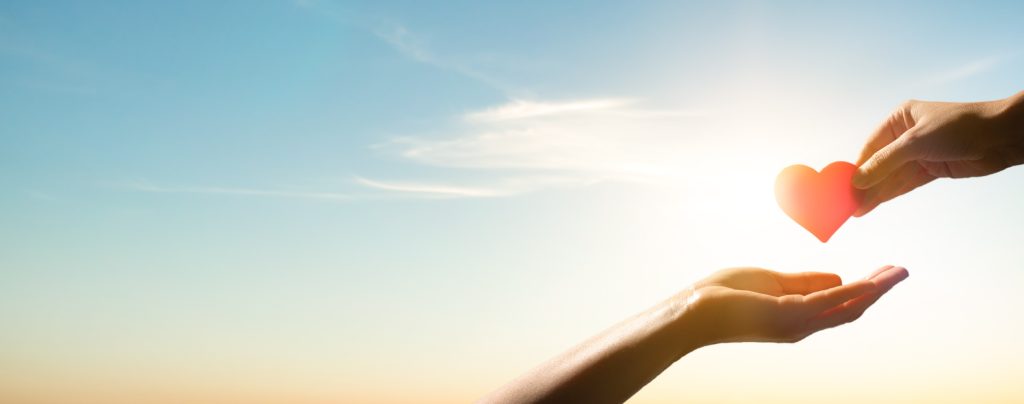 Two hands reaching towards each other in a heart-giving gesture against a bright sky.