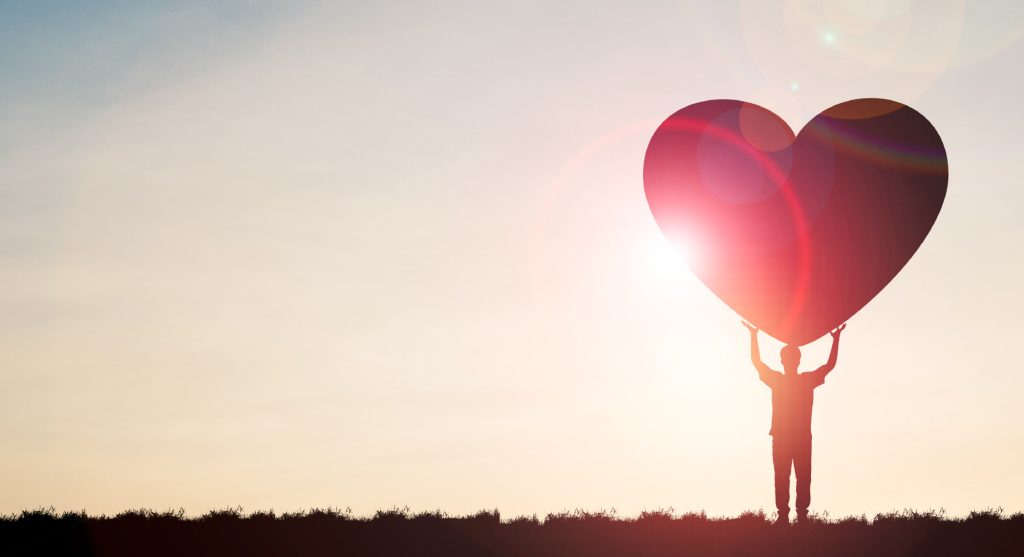 Silhouette of a person holding up a large heart shape against the sky.