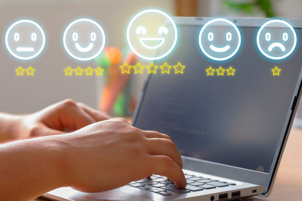 Close-up of hands typing on a laptop with a row of neon smiley face icons above the screen, each with a different expression representing various levels of satisfaction.