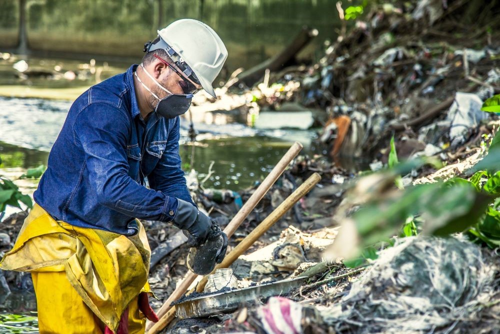 Worker wearing protective gear, including a hard hat, gloves, and face mask, cleaning a polluted area near a body of water.