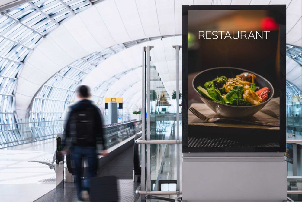 Digital sign in an airport terminal displaying a restaurant advertisement with a bowl of food, while a traveler walks past with luggage.