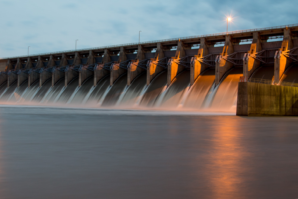 Hydroelectric dam releasing water through multiple spillway gates at dusk, with warm lights reflecting on the surface.