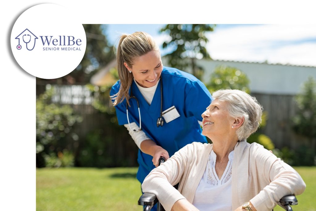 Smiling nurse in blue scrubs caring for an older woman in a wheelchair outdoors, with the WellBe Senior Medical logo in the corner.