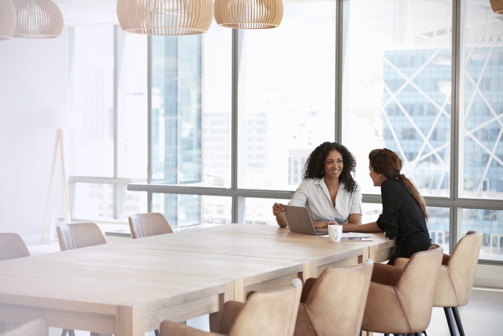 Two women sitting at a long conference table in a bright modern office, talking and working on a laptop with city views in the background.
