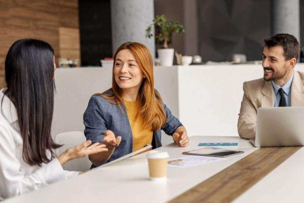 Three colleagues sit together at a table, smiling and talking during a casual meeting with papers, a laptop, and a coffee cup in front of them.
