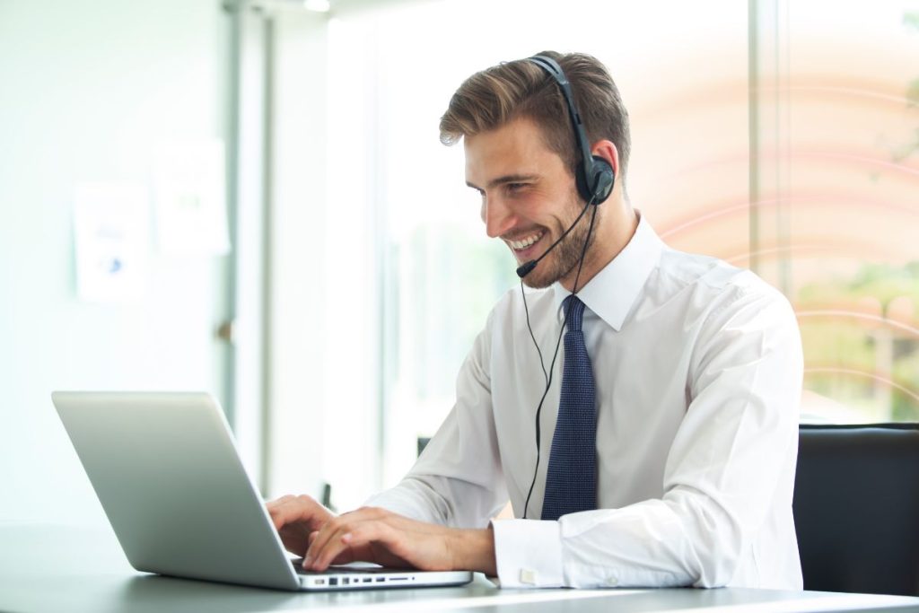 Customer service agent wearing a headset and tie, working on a laptop in a bright office environment.