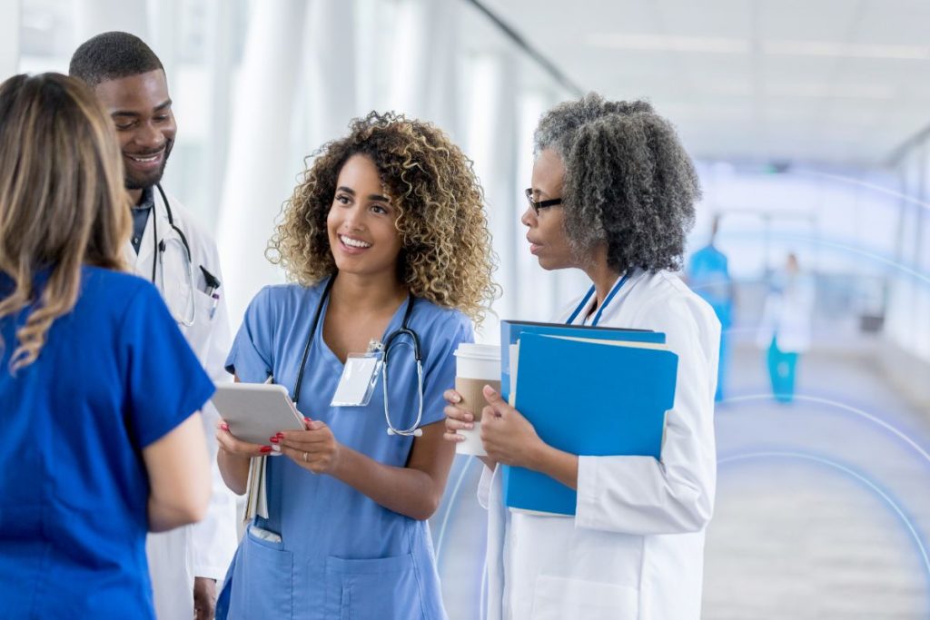 Healthcare clinicians collaborating in a hospital corridor, reviewing patient information on a tablet and discussing care coordination.