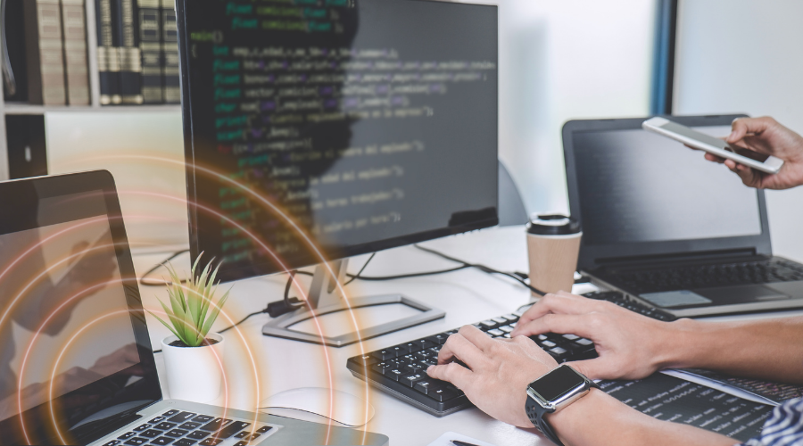 hands typing on a keyboard with a monitor and someone holding a laptop